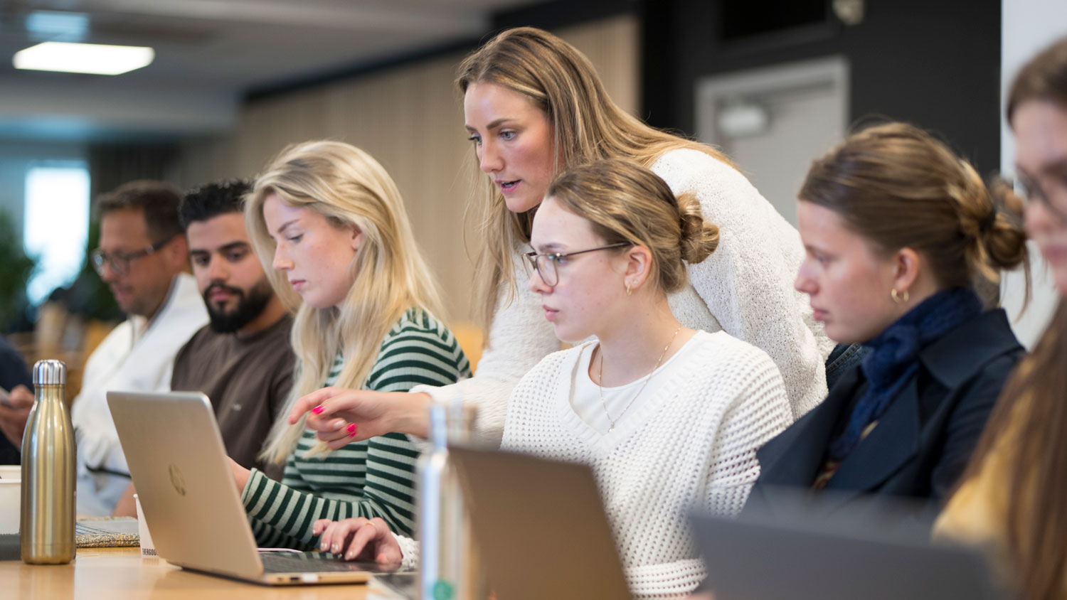 En kvinne står og peker på en bærbar PC-skjerm for å veilede en kollega i et lyst kontormiljø. Flere kolleger sitter ved siden av hverandre og arbeider konsentrert på sine laptoper.